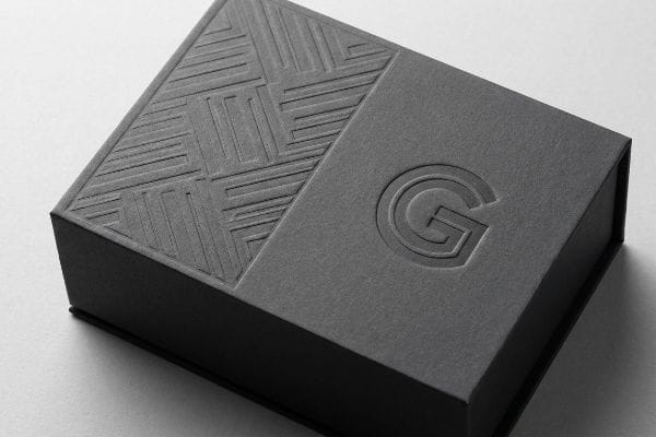 A dark grey packaging box features an embossed geometric pattern and a circular G logo, highlighting premium tactile finishes.
