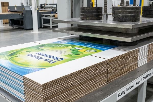 Heavy circular weights on metal plates press down on stacks of litho-laminated corrugated panels, featuring an upside-down 