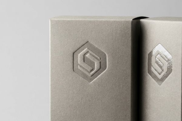Textured light grey packaging features a debossed hexagonal logo, with one showing a matte finish and the other a reflective spot UV effect, highlighting the physical depth.
