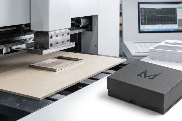 Steel rule die on a debossing machine presses into a cardboard sheet, next to a black debossed box with an 'M' logo.
