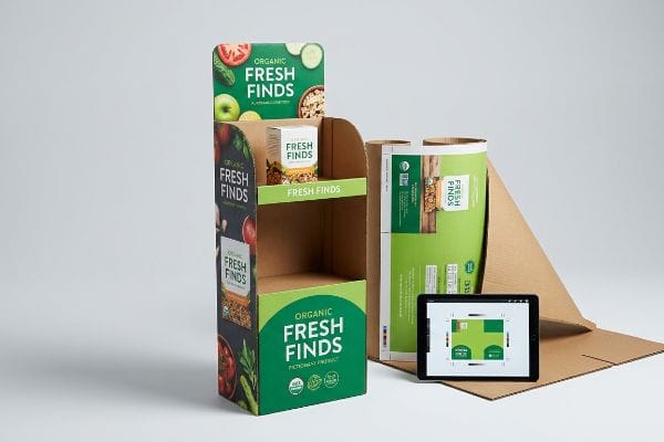 Customizing an Organic Fresh Finds cardboard display stand, featuring a finished unit, rolled printed artwork, and digital design on a tablet.