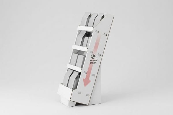 White corrugated cardboard display, loaded with gray products, illustrates 'Center of Gravity' with a red arrow, highlighting tip-over physics.