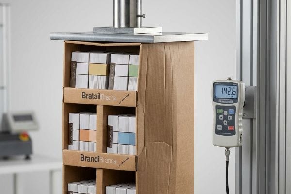 Corrugated cardboard display for Bratall and Brandi undergoing a top-load compression test, with a digital force gauge showing 142.6 lbs, evaluating structural integrity.