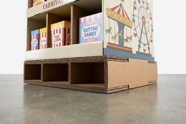 A carnival-themed cardboard display rack with Pop Corn and Cotton Candy boxes, showing its double-wall corrugated base on a concrete floor.