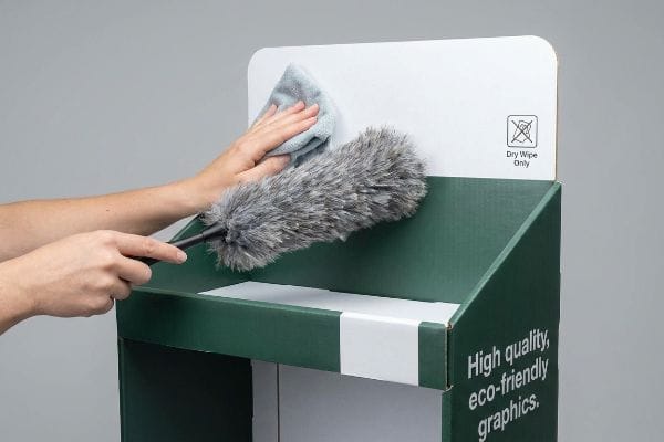 Hands dry wiping a green and white cardboard display with a static duster and microfiber cloth, highlighting 'Dry Wipe Only' and 'High Quality, Eco-Friendly Graphics'.