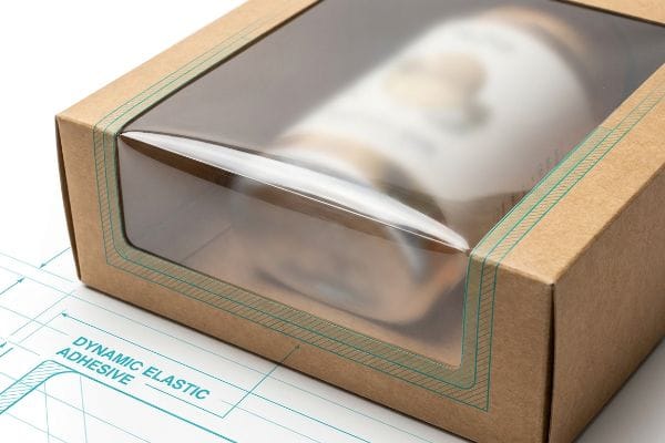 Brown kraft cardboard showcase with a clear viewing window, highlighting the Dynamic Elastic Adhesive on a technical blueprint.