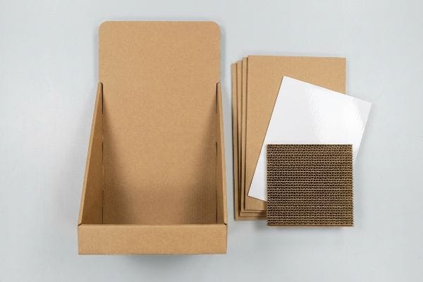 An overhead view shows unassembled brown corrugated PDQ display components, including flat sheets, a glossy white clay-coated top sheet, and exposed E-flute.
