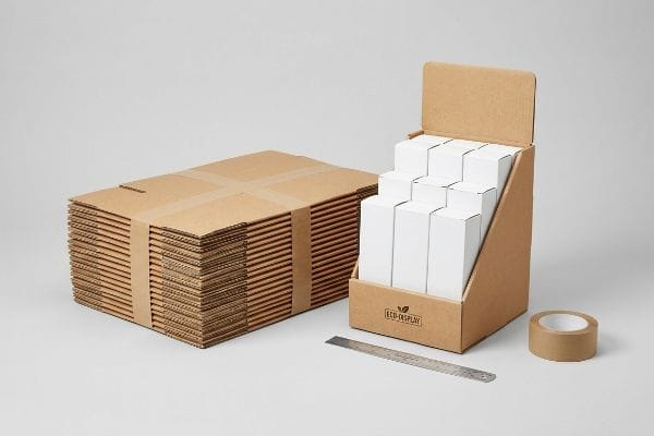 Solución de caja de exhibición de cartón En una fotografía de estudio se muestran cajas de cartón desmontadas y una caja de venta al por menor Eco-Display ya montada, llena de productos blancos, junto con una regla y cinta adhesiva.