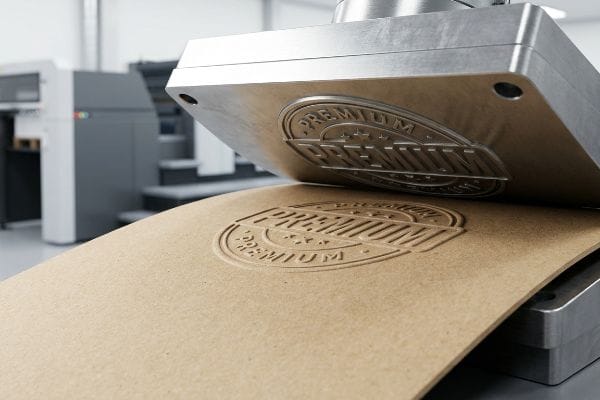 An embossing machine presses a metal die, creating a raised Premium logo on brown paperboard, showcasing tactile packaging.