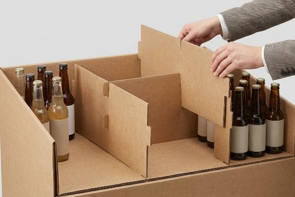 Kraft paperboard modular display box with friction-fit interlocking dividers being inserted, segmenting amber glass bottles for merchandising.