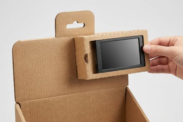 A hand removes a black electronic device from a brown corrugated cardboard display with a friction-lock pop-out housing, enabling tool-free e-waste separation.