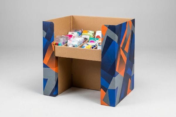 Présentoir en carton ondulé de type bac de rangement orné de motifs géométriques bleu vif, orange et gris, rempli de marchandises d'achat impulsif assorties dans un plateau compartimenté.
