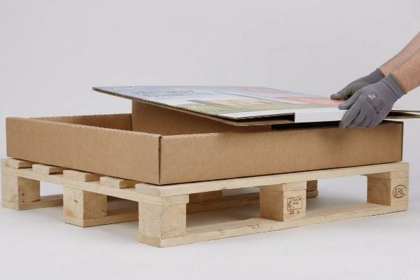 Gloved hands place a printed corrugated lid onto a brown half pallet display box, resting on a wooden EPAL pallet.
