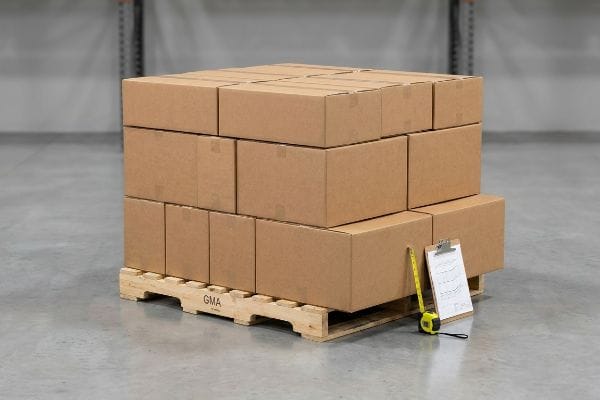 Brown cardboard master cartons stacked on a wooden GMA pallet, with a yellow measuring tape and clipboard for dimension checks.