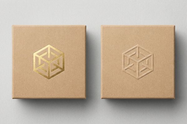 Comparing two kraft paper boxes, one with a shiny gold foil stamped geometric logo and the other with a subtle embossed version.