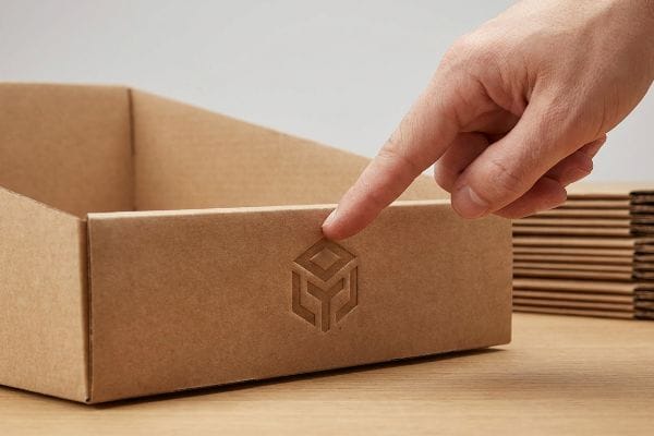 Brown corrugated cardboard box featuring a debossed geometric logo, emphasized by a finger pointing to its tactile finish.