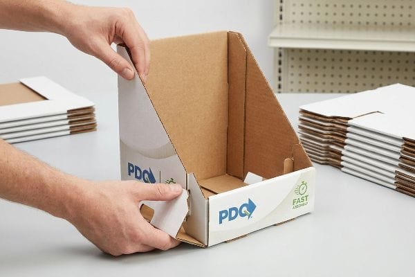 A person's hands assemble a PDQ corrugated retail display, featuring white exterior branding with 'PDQ' and 'Fast Assembly' logos, utilizing interlocking tabs.