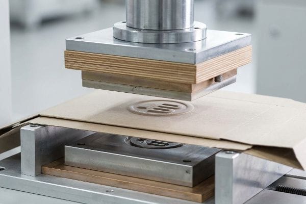 A metal die applies pressure to kraft cardboard, creating a crisp embossed logo for retail packaging.
