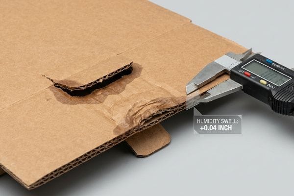 Corrugated B-flute cardboard thickness measured by a digital caliper, showing a Humidity Swell of +0.04 Inch due to moisture absorption.