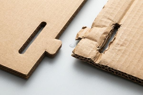 Corrugated paperboard with a clean locking tab next to a torn, humidity-swollen slot and water droplet, showing structural failure.