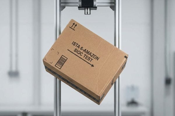 Brown cardboard box, labeled 'Ista 6-Amazon Sioc Test' with a barcode, suspended in a metal testing rig for packaging drop test.