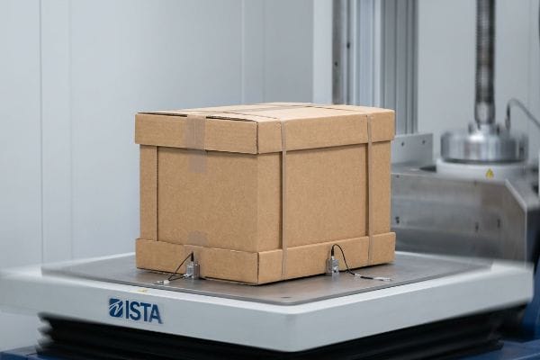 Brown corrugated cardboard box undergoing ISTA transit vibration testing with attached sensors on a specialized machine.
