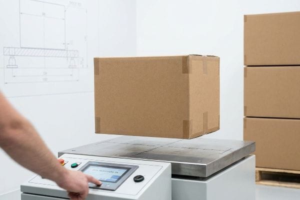 A person's hand operates a testing machine performing Ista Procedure 3a on a floating brown cardboard box.
