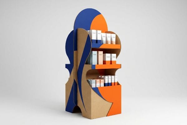 Kraft cardboard retail display featuring bold blue and orange die-cut geometries, showcasing cosmetic products on three shelves.