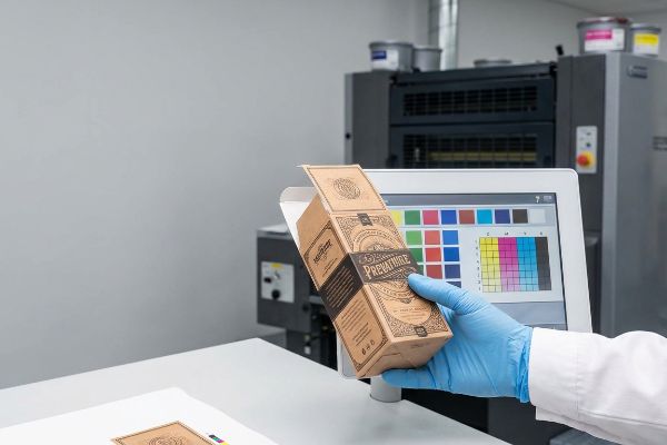 Brown kraft paper 'Prevatune' packaging box held by a gloved hand, inspected against a CMYK color calibration monitor and printing press.
