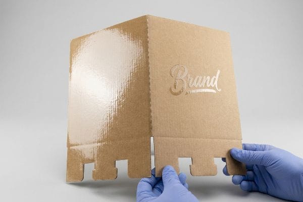 Brown corrugated cardboard display featuring glossy UV coating on one panel and matte interlocking tabs, held by hands in blue gloves.