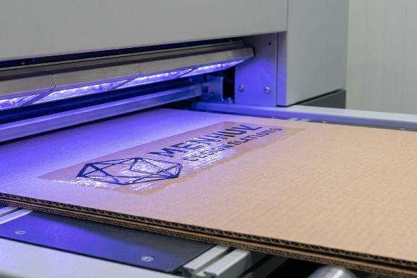 Corrugated cardboard with Menhul Design Solutions logo receiving a raised spot UV gloss finish under blue ultraviolet light.