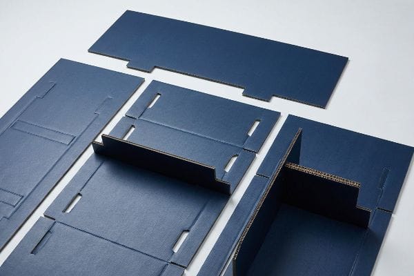 Engineered dark blue micro-fluted cardboard components for a flat-pack POP display are shown in various stages of assembly.
