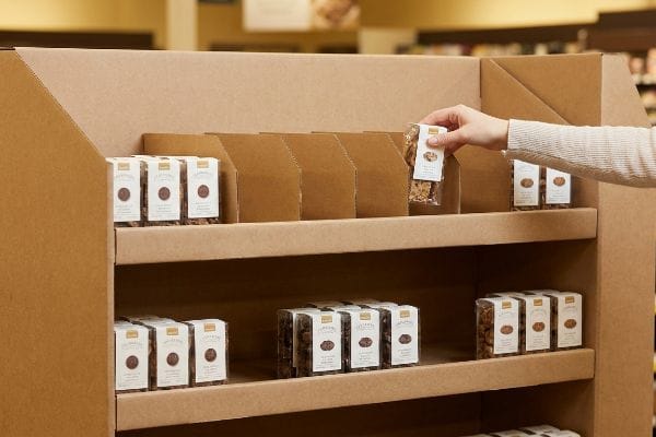 Corrugated cardboard retail display with modular dividers, showcasing packaged granola or nuts. A hand places a product, demonstrating easy restocking.