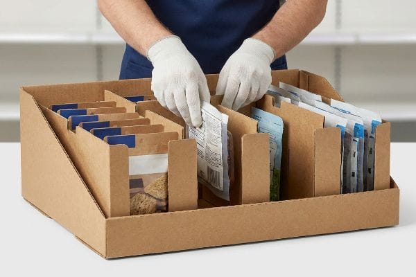 Restocking Retail Display Tray Brown corrugated cardboard display tray with modular dividers, being restocked by gloved hands placing product pouches for efficient shelf replenishment.