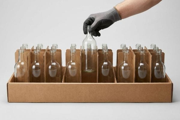 Brown corrugated box with modular dividers, a gloved hand placing a clear glass bottle, illustrating packing clearance for merchandising.