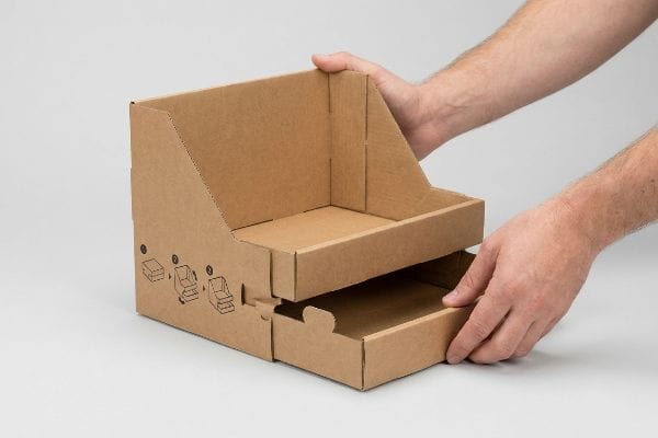 Natural brown Kraft cardboard modular display box with two tiers, featuring pre-glued assembly diagrams and hands demonstrating setup.