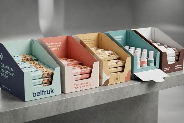 Corrugated cardboard shelf-ready displays showcase Belfruk snack bars, small cosmetic bottles, and packaged goods in multi-colored, branded trays on a grey counter.