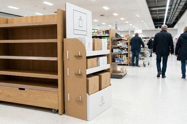 Dans une allée d'un magasin, on trouve un présentoir PLV en carton avec des boîtes de produits Nova Scent en kraft et blanches, à côté d'étagères en bois vides.