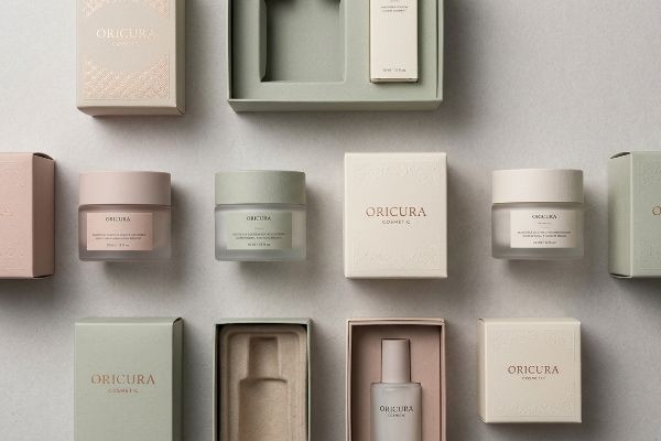 An overhead view displays Oricura Cosmetic skincare products and their elegant pastel packaging, including jars, bottles, and boxes.