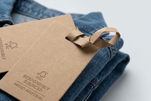 Kraft paper hang tags with FSC Responsibly Sourced Mono-Material text and an origami-style paper lock attached to blue denim.