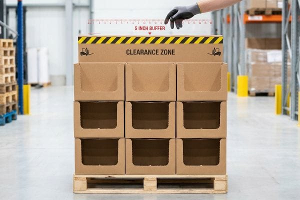 A hand measures a 5 Inch Buffer above brown cardboard display boxes on a pallet, indicating a 'Clearance Zone'.