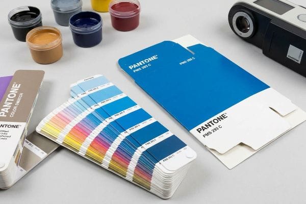 A collection of Pantone color tools, including a fan-shaped color swatch, two PMS 293 C blue cards, small jars of various colored inks, and a colorimeter.