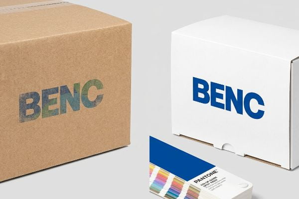 Raw cardboard box with faded Benc logo from process inks, contrasted with a white box featuring a vibrant blue Benc logo from spot color, beside a Pantone Color Guide.