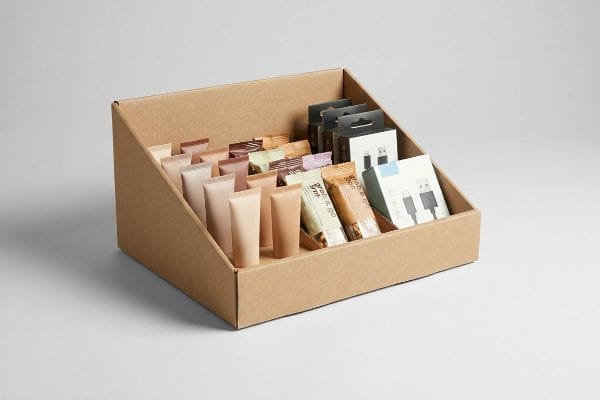 A brown cardboard PDQ display tray filled with lightweight cosmetic tubes, Grab & Go snacks, and small electronics like USB cables.