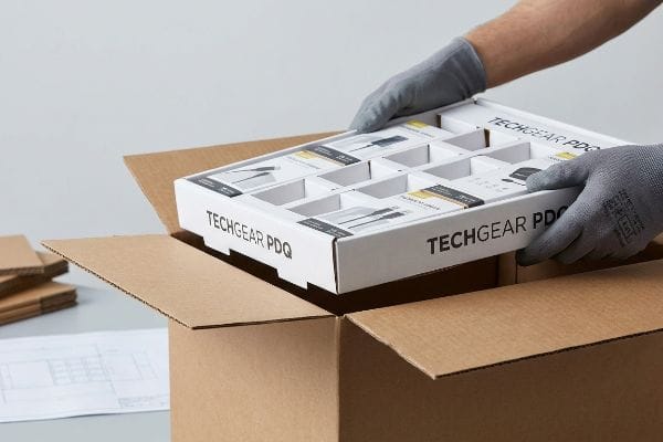 White Techgear PDQ display tray, pre-filled with products, is carefully placed by gloved hands into a brown corrugated shipping box.