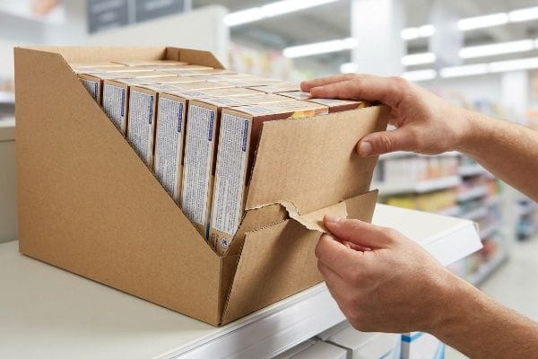 Brown corrugated PDQ display tray opened by hands, tearing front panel to reveal product boxes on a retail shelf.