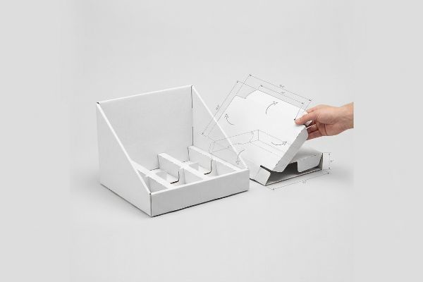 White corrugated PDQ display and a hand assembling a die-cut retail-ready tray, showing fold lines and engineering measurements.