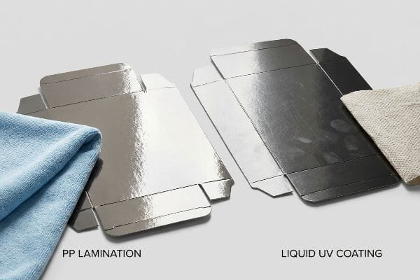 Shiny Pp Lamination cardboard box with a blue microfiber cloth, contrasted with a smudged Liquid Uv Coating box and a beige paper towel, showing finish durability.