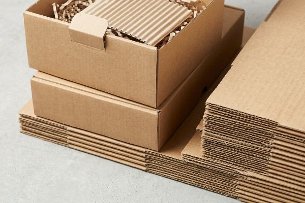 Stacked brown corrugated cardboard boxes, one open with shredded paper filler, beside flat raw testliner sheets.