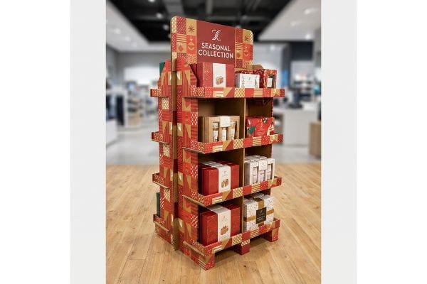 Holiday Packaging Display Stand A multi-tiered corrugated cardboard display, adorned with red and gold festive patterns, showcases Seasonal Collection gift sets in a retail store.